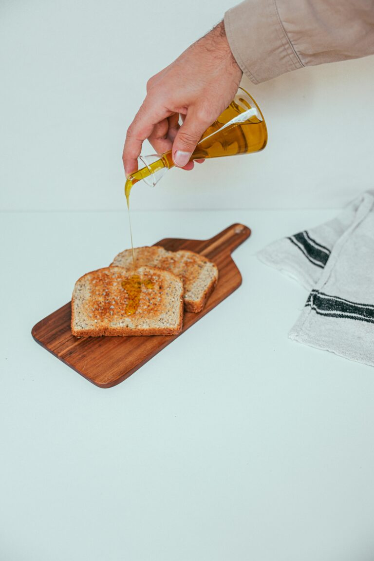 Hand pouring olive oil on wheat toast slices on wooden board.